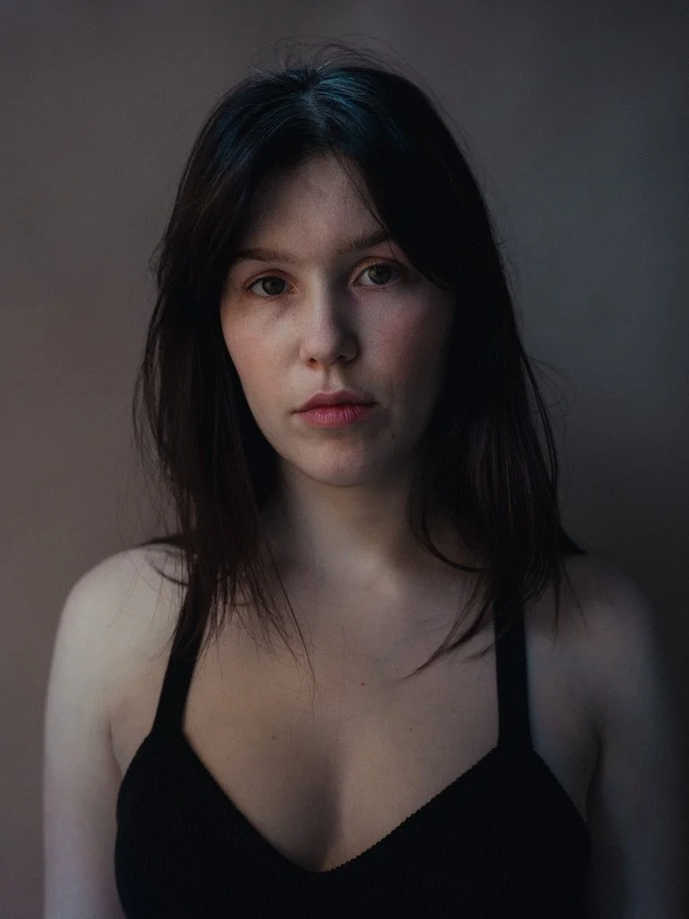 A young woman with dark hair and light skin, wearing a black tank top, looking directly at the camera against a plain background.