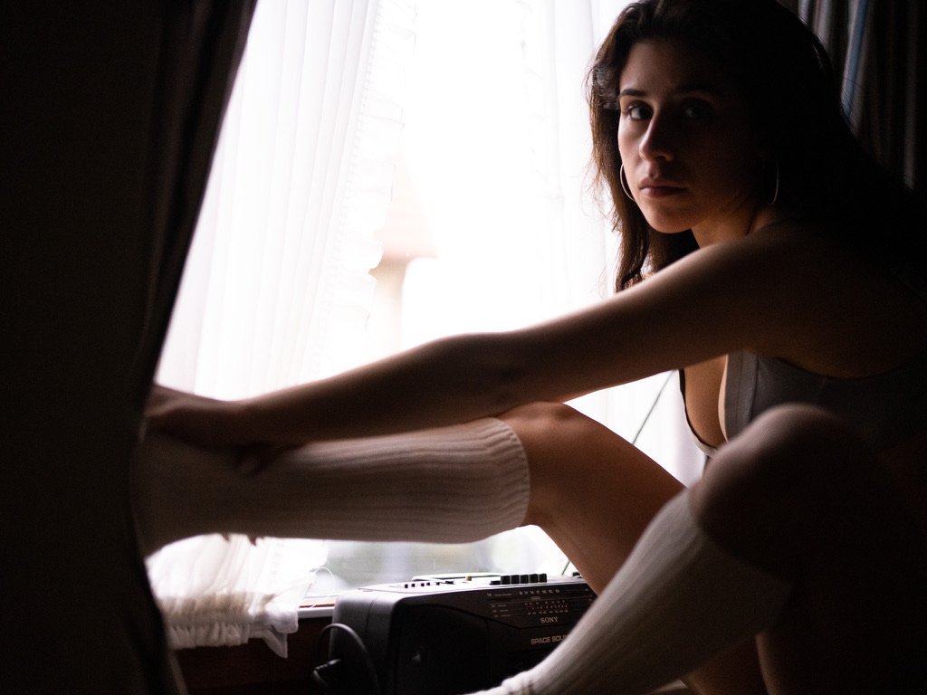 Woman sitting by a window with sheer curtains, stretching her leg and looking towards the camera, wearing knee-high socks.