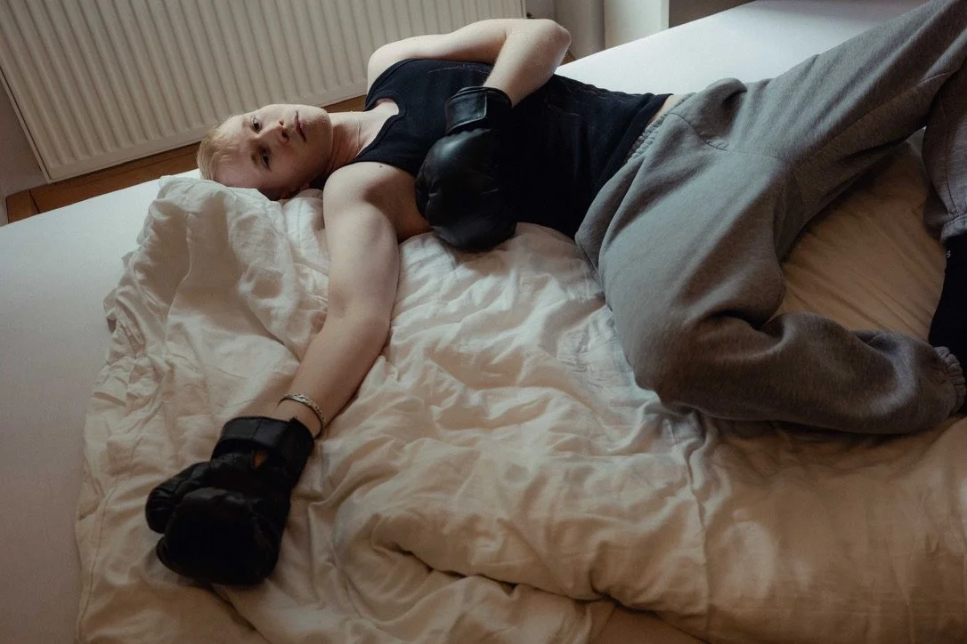 A young man with blonde hair lying on a bed with white sheets, wearing a black tank top and gray sweatpants, with a black dog resting on his chest.