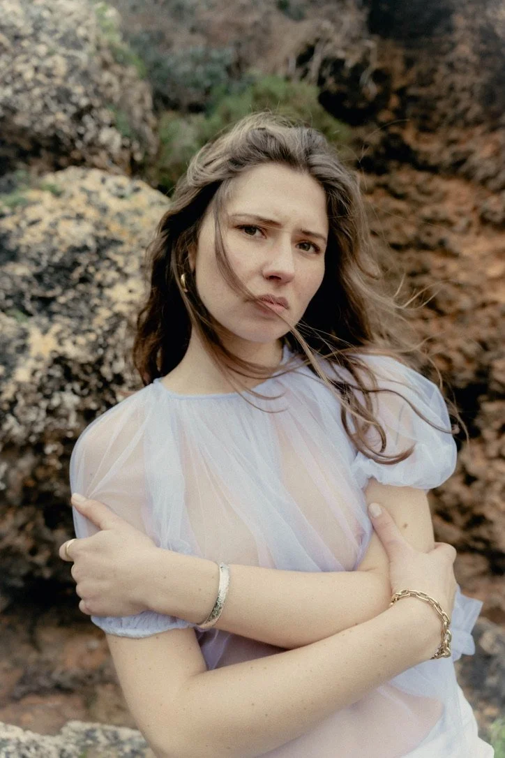 A young woman with long brown hair standing outdoors in front of rocky terrain, wearing a sheer white blouse with puffy sleeves, looking into the camera with a serious expression, arms crossed.