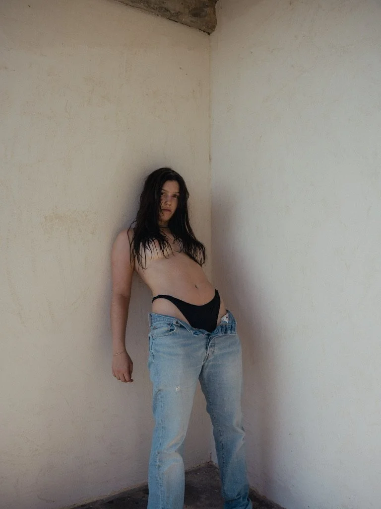 A woman with long dark hair standing topless in a corner of a room, wearing loose jeans and black underwear, with a neutral expression.