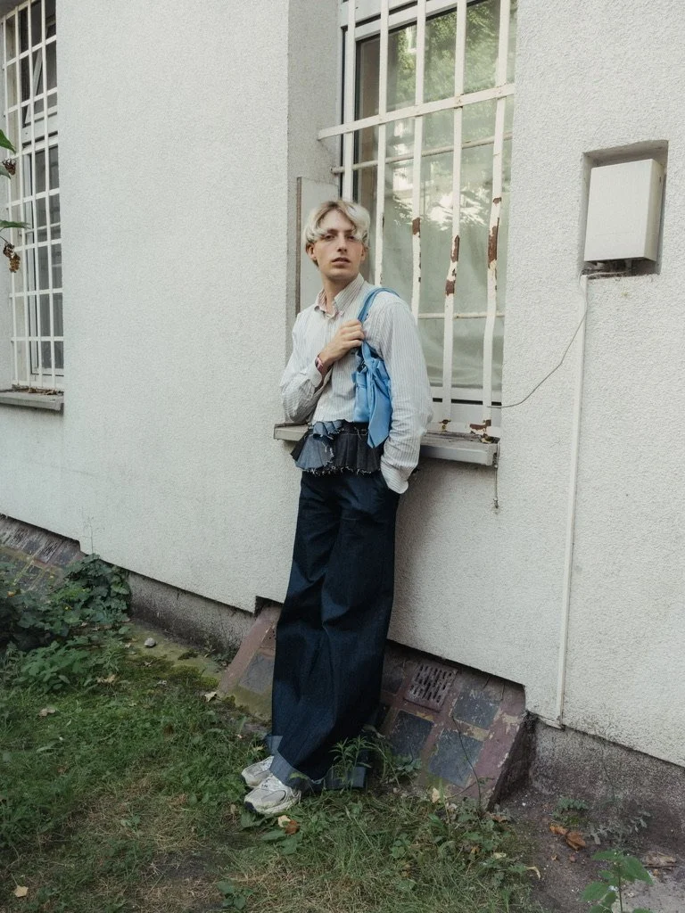 A young person with short light-colored hair, wearing a light striped shirt and large black pants, standing outside next to a white wall with a window that has metal bars, carrying a light blue backpack.