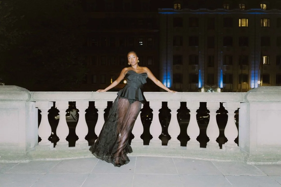 A woman in a stylish evening outfit standing on a balcony at night, with a city building lit in the background.