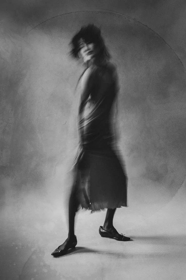 A black and white photo of a woman with blurry motion, wearing a dress and high heels, standing against a plain background.