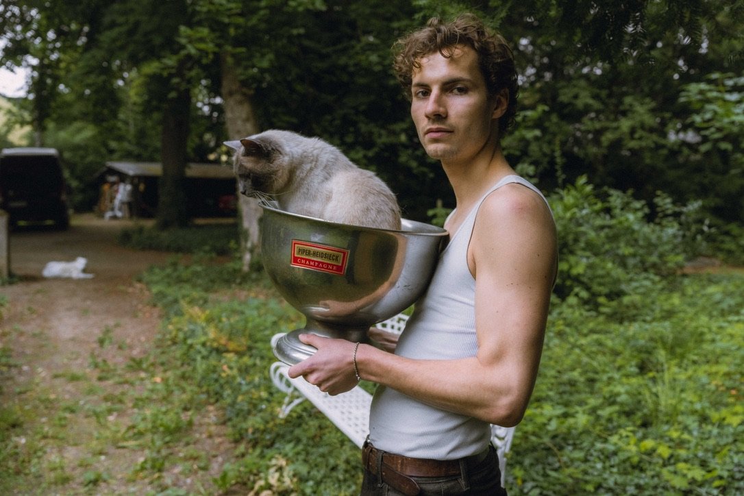 A young man with curly hair wearing a sleeveless shirt holds a large chrome bowl with a gray cat inside. They are outdoors in a wooded area.