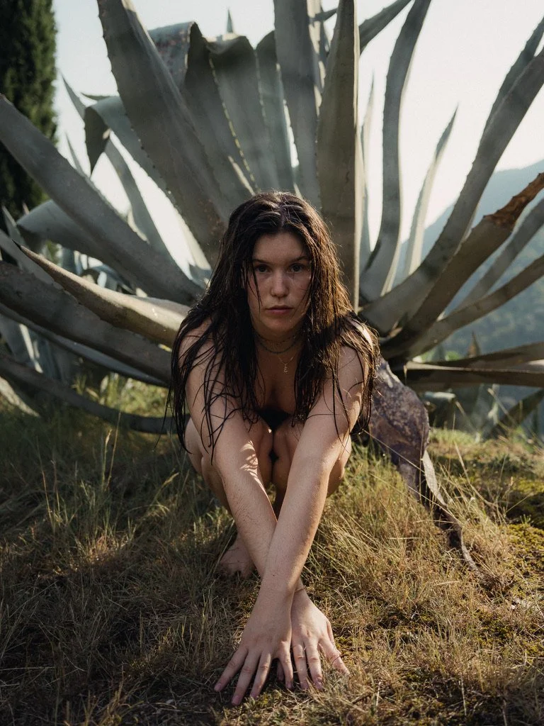Young woman with wet hair crouching on the ground in front of a large agave plant, looking intently at the camera.