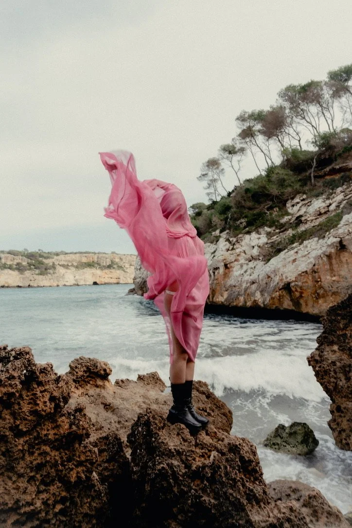 Person standing on rocks by the ocean with pink fabric flowing around them and coastal cliffs in the background.