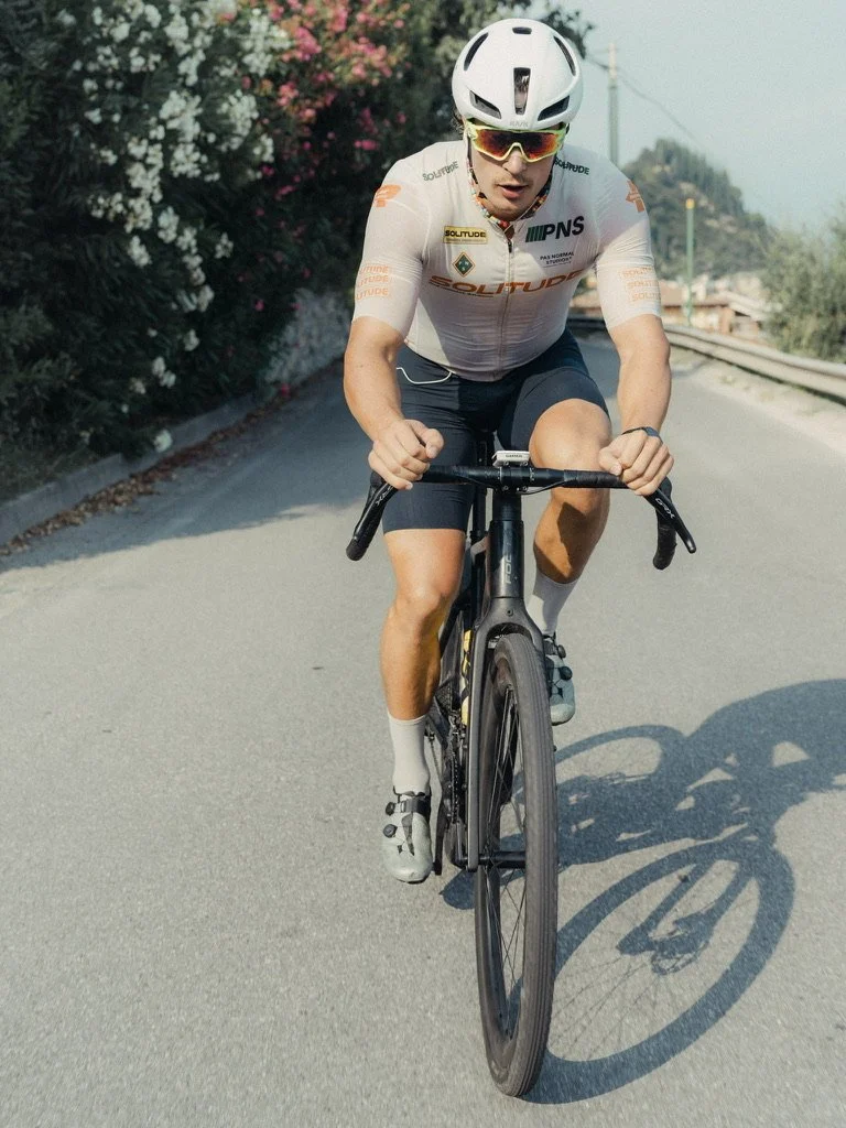 A man riding a black road bike uphill on a sunny day, wearing a white helmet, sunglasses, a white cycling jersey, black shorts, white socks, and cycling shoes, with trees and flowers in the background.