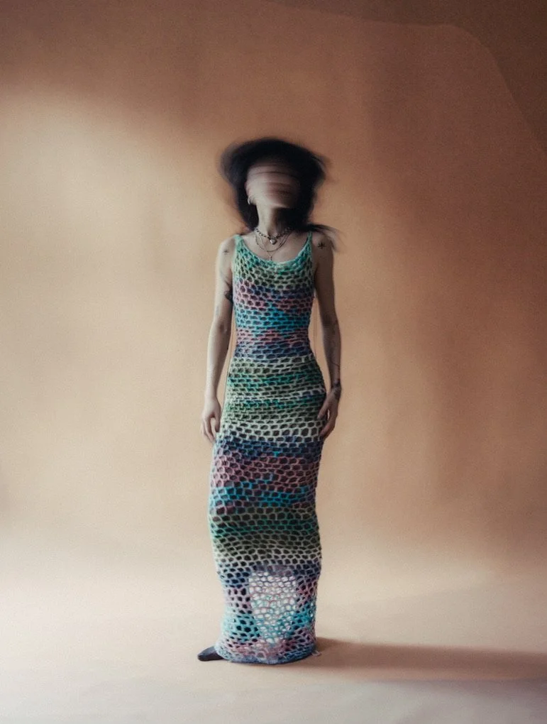 A person with long, dark hair wearing a colorful, open-knit sleeveless dress stands against a plain beige background, with their face blurred.