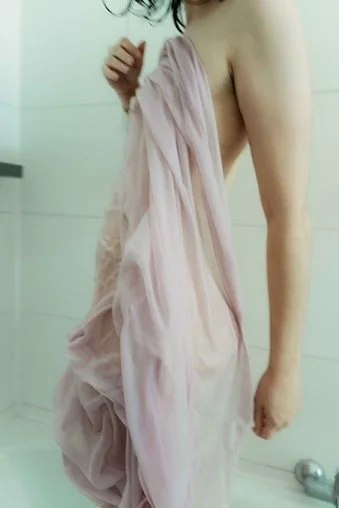 A person wrapped in a light pink towel in a bathroom with white tiled walls.