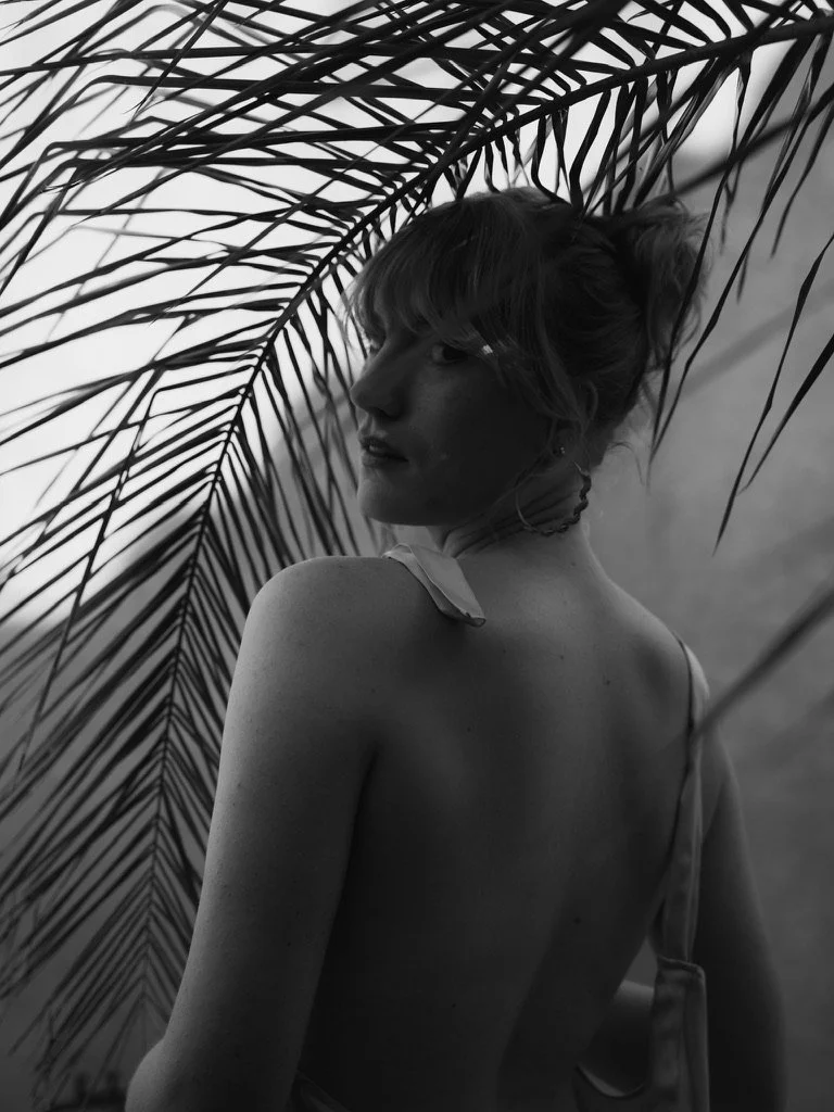 A black and white photo of a woman with short hair, turned slightly away, surrounded by palm fronds.