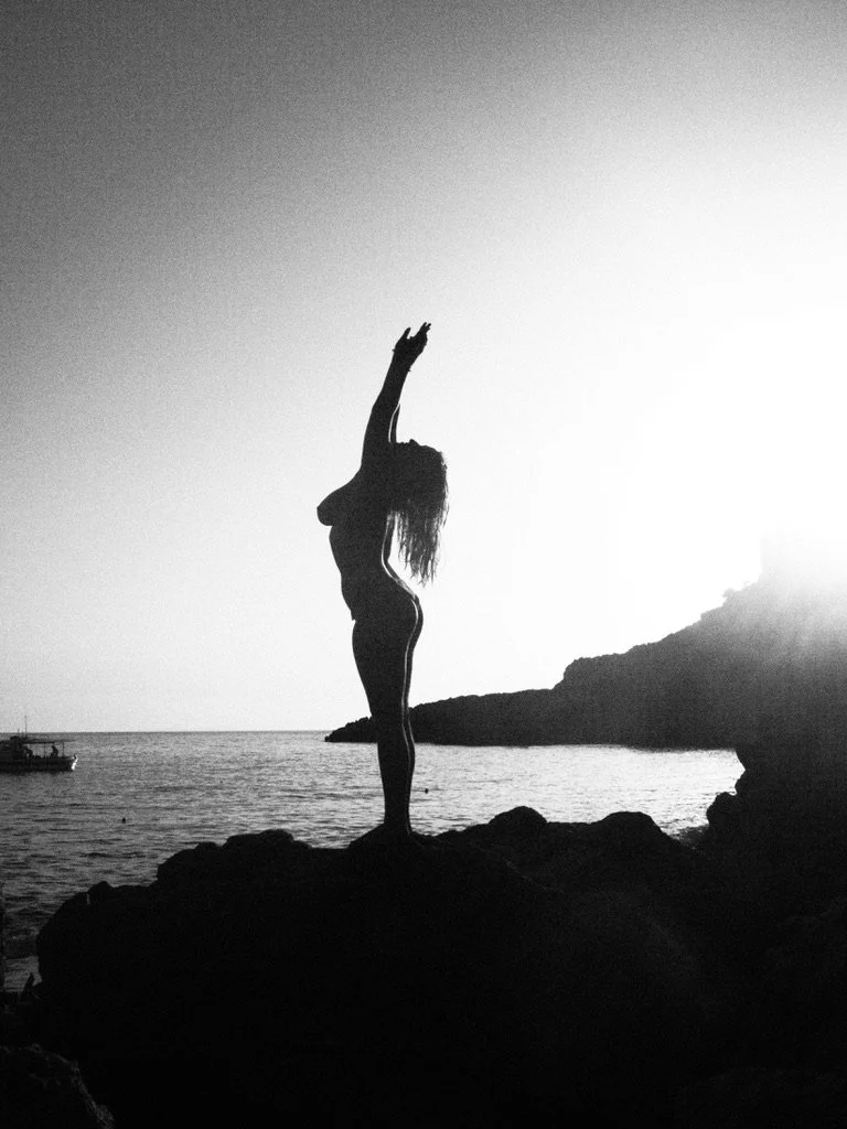 Silhouette of a woman standing on rocks by the water with her arms raised above her head at sunset.