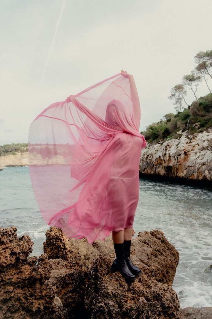 Person in a flowing pink dress and black boots standing on a rocky shoreline during daytime, holding the dress up with one arm, with a body of water and rocky cliffs in the background.