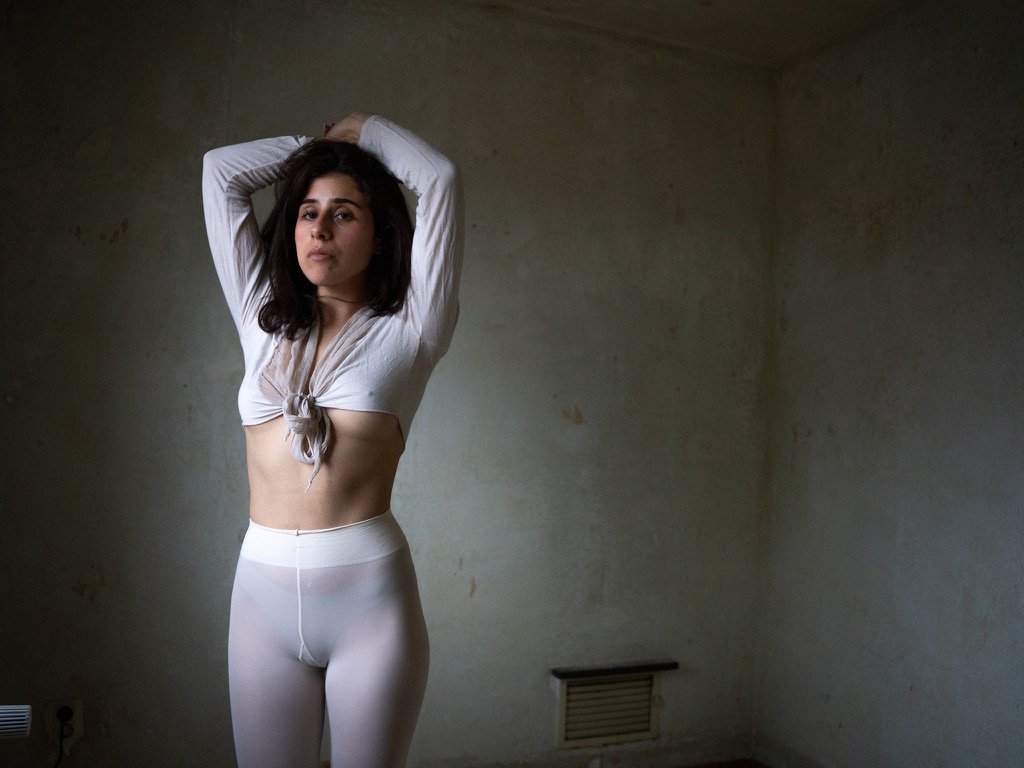 A woman standing with her arms raised and hands behind her head in a minimally furnished room with plain walls.