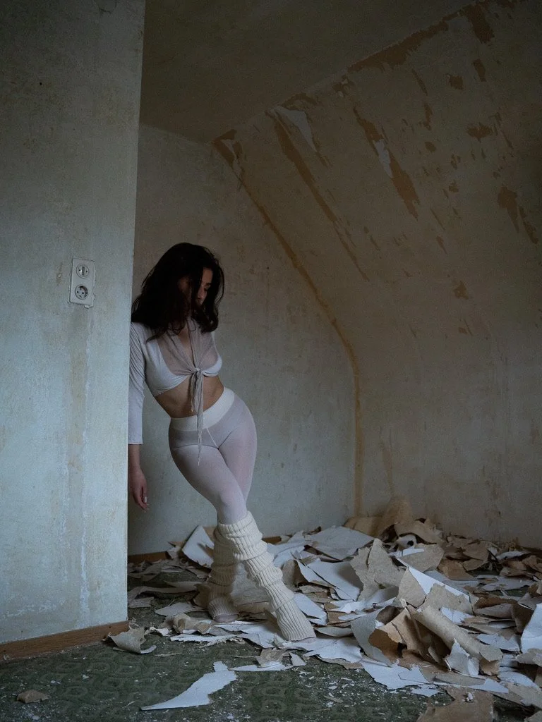 A woman standing in an unfinished, dilapidated room with torn wallpaper and scattered paper debris on the floor.
