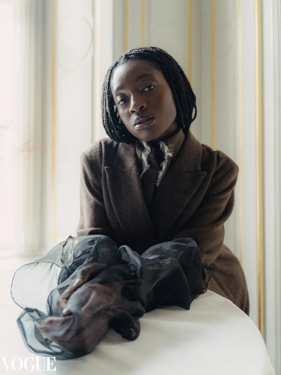 A woman sitting at a table in an elegant room with yellow and white striped wallpaper, wearing a brown blazer and holding a black sheer fabric, gazing at the camera in a Vogue photoshoot.