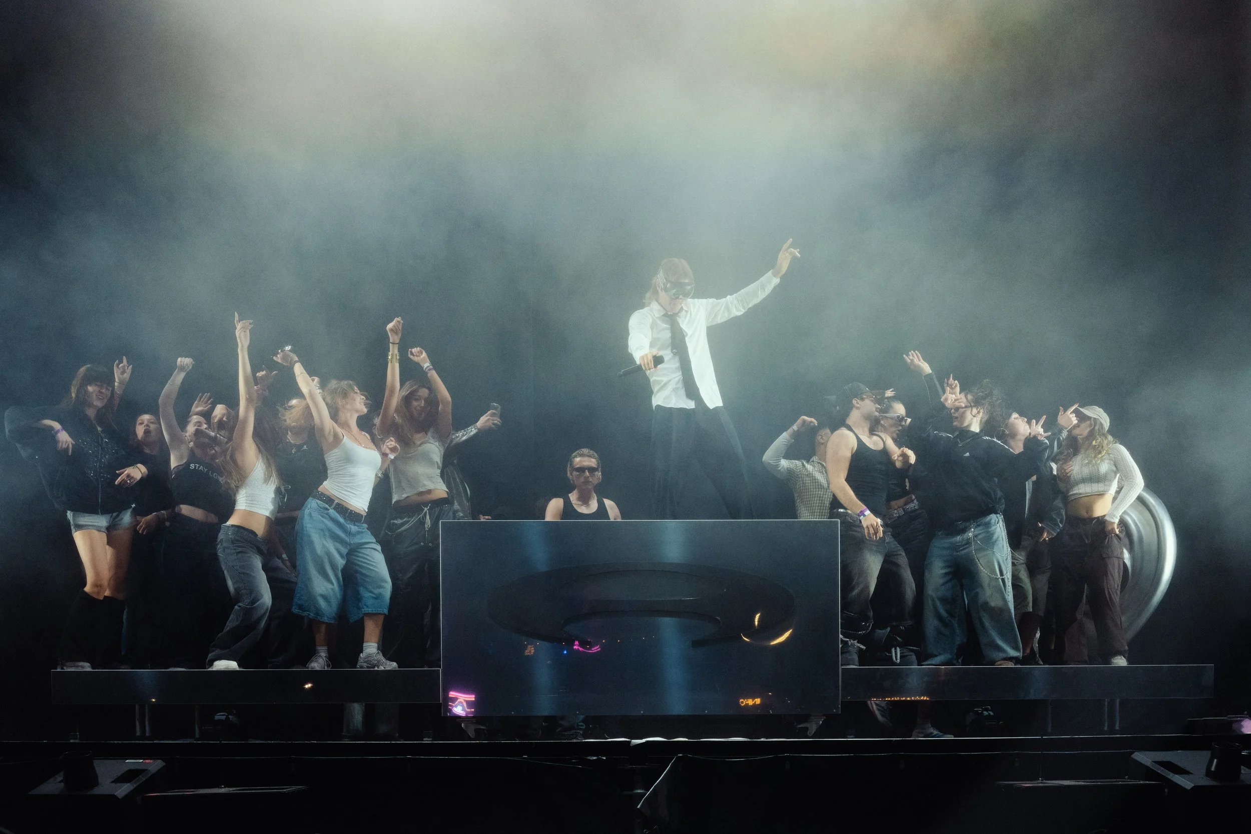 A DJ in a white shirt, black tie, and headphones stands on a platform surrounded by young people dancing and enjoying music at a concert or nightclub with smoky atmosphere and dim lighting.