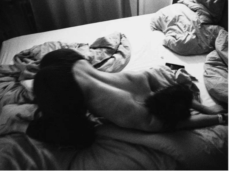 A person is lying on a bed with their body stretched out, accompanied by a dog resting beside them. The bed has rumpled sheets and pillows, and a remote control is on the bed. The image is in black and white.