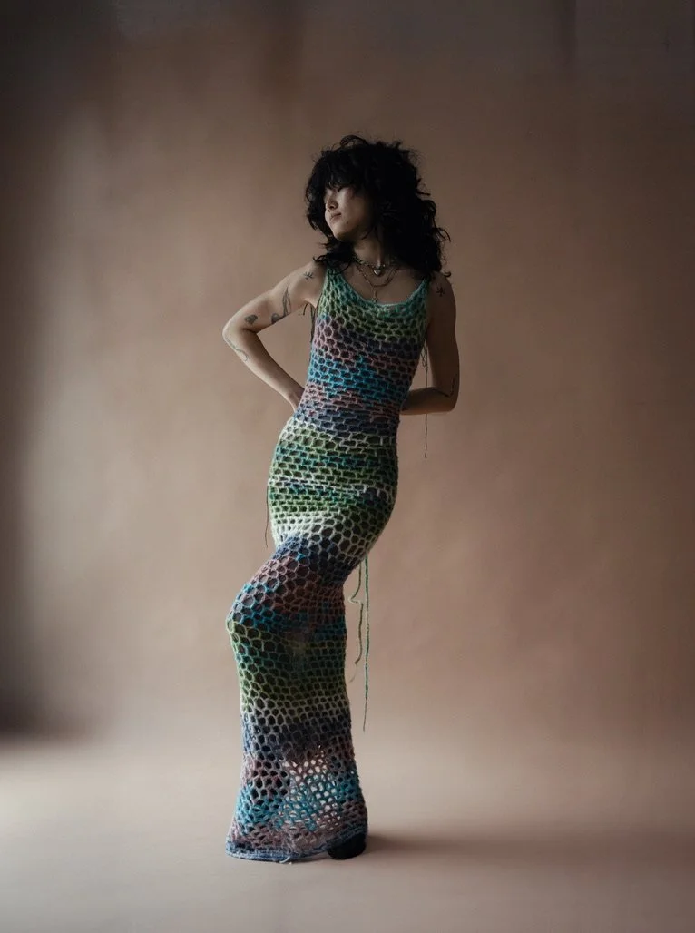 A woman with dark curly hair and tattoos stands in front of a neutral background, wearing a colorful, textured, full-length dress with a fishnet pattern.