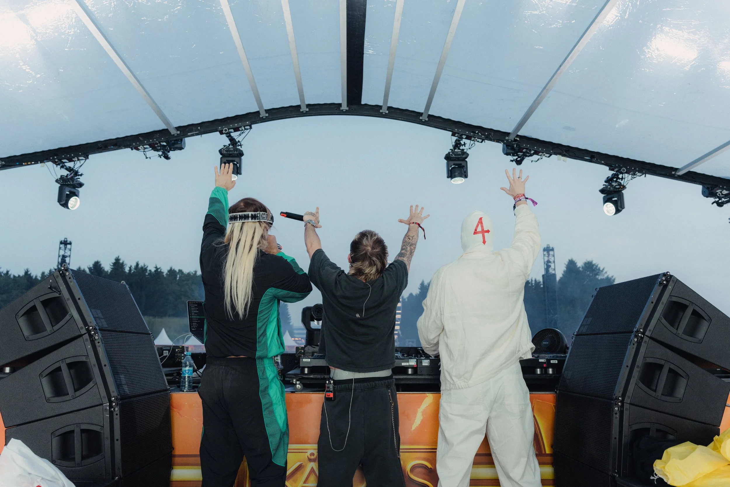 Four people on a stage at an outdoor music festival, with their hands in the air. Two of the individuals are DJ gear users, with one wearing a white hoodie with a red number 4 on the back, another with long blonde hair and headphones, another with short hair and tattooed arm, and the last person partially visible.