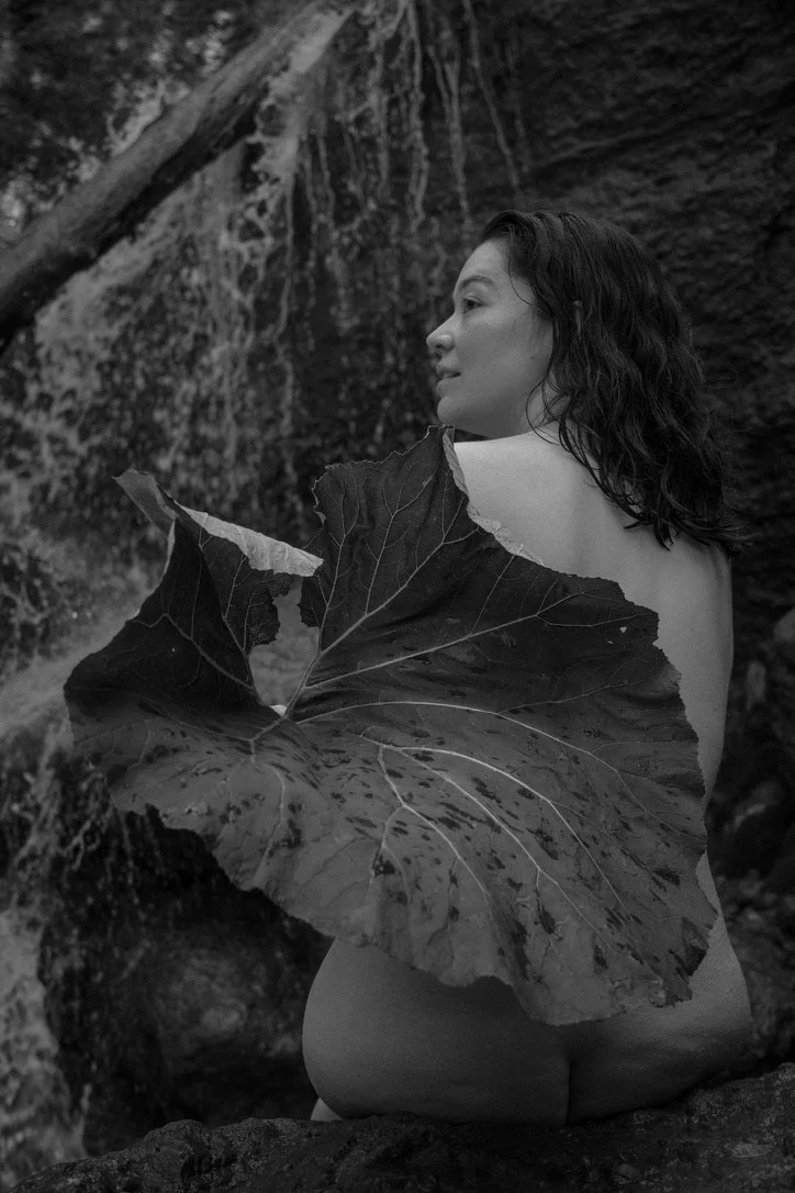 Black and white photo of a woman with shoulder-length hair sitting outdoors near rocks and a waterfall, with a large leaf covering her back.