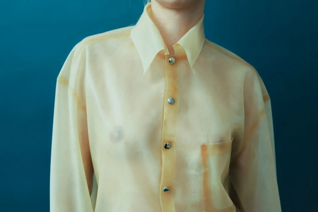 Close-up of a person wearing a shiny, pale yellow button-up shirt with a pocket on the left side, standing against a dark teal background.