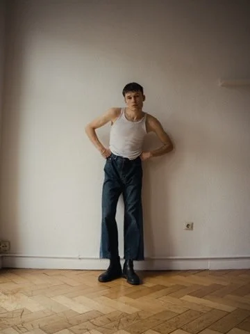 Young man standing against a plain white wall with hands on hips, wearing a white tank top, high-waisted blue pants, and black shoes.