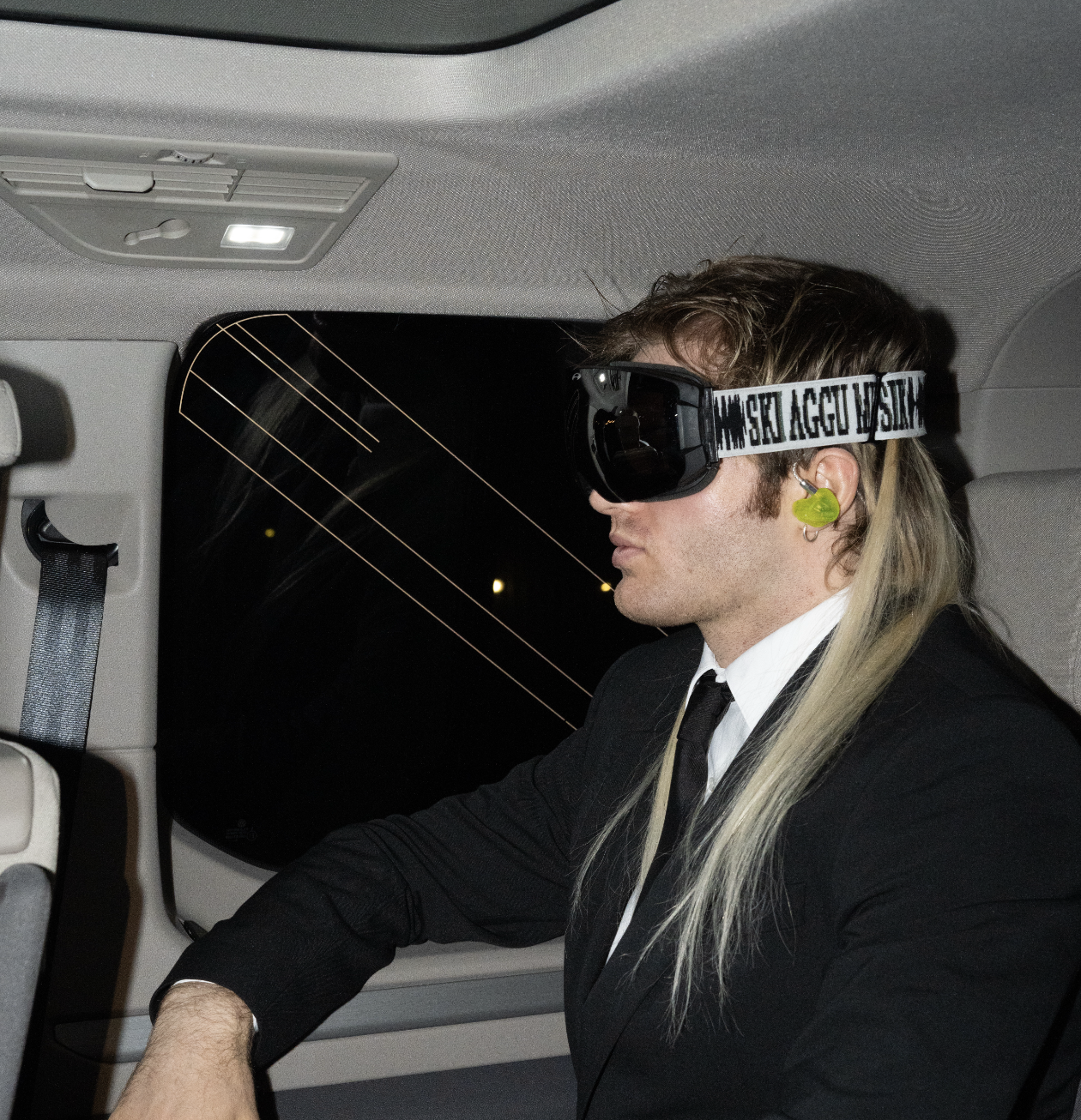 A man with long blonde hair wearing a black suit, white shirt, and black tie, sitting in the back seat of a vehicle at night. He is wearing large black goggles with printed text on the strap and green earplugs.