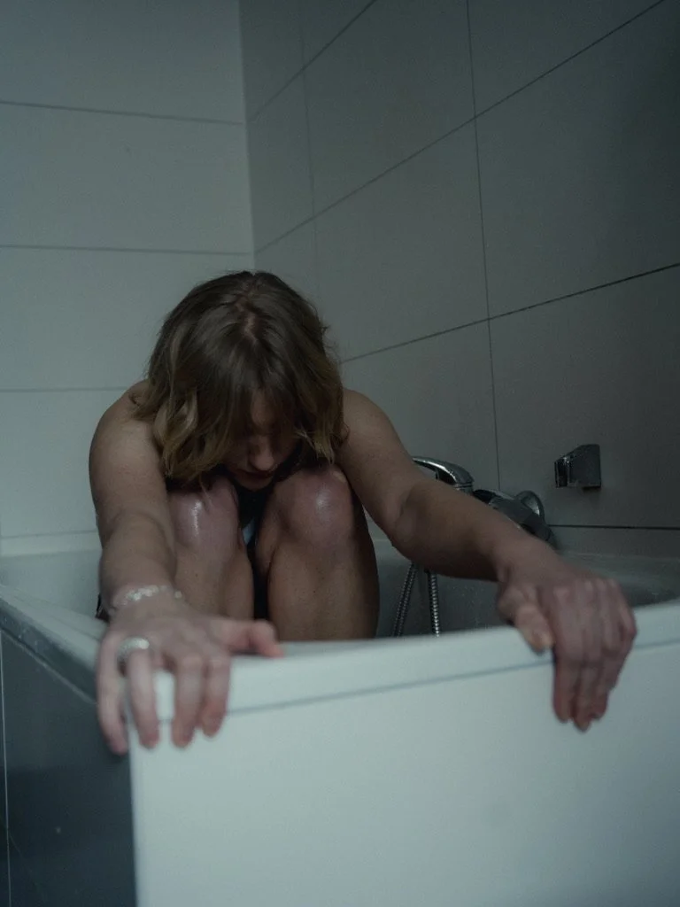 A woman in a bathtub holding onto the sides with her hands, her head bowed and hair covering her face.
