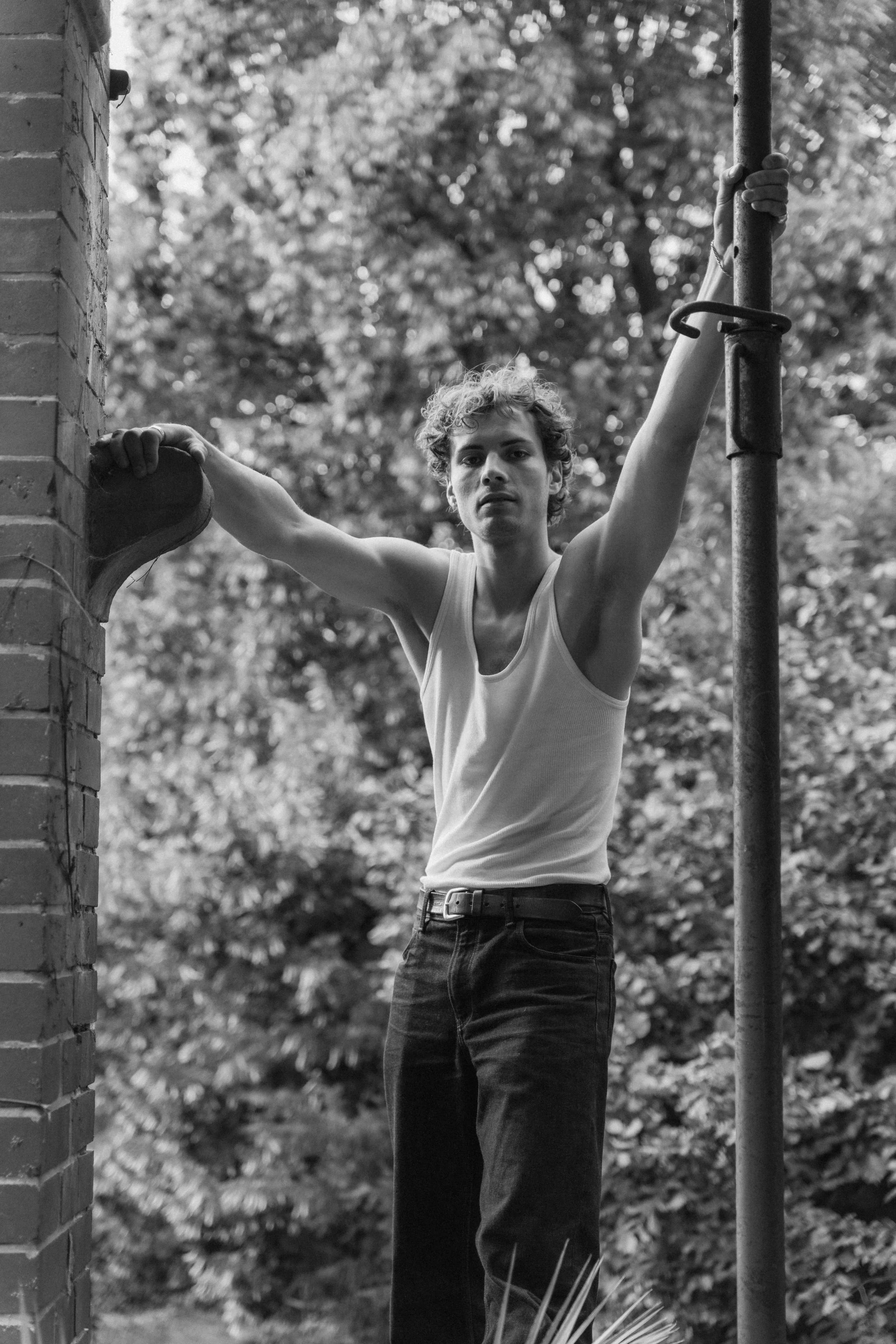 A young man with curly hair in a sleeveless shirt is outdoors, hanging onto a vertical pipe with one hand and holding a shoe with the other, with trees and foliage in the background.