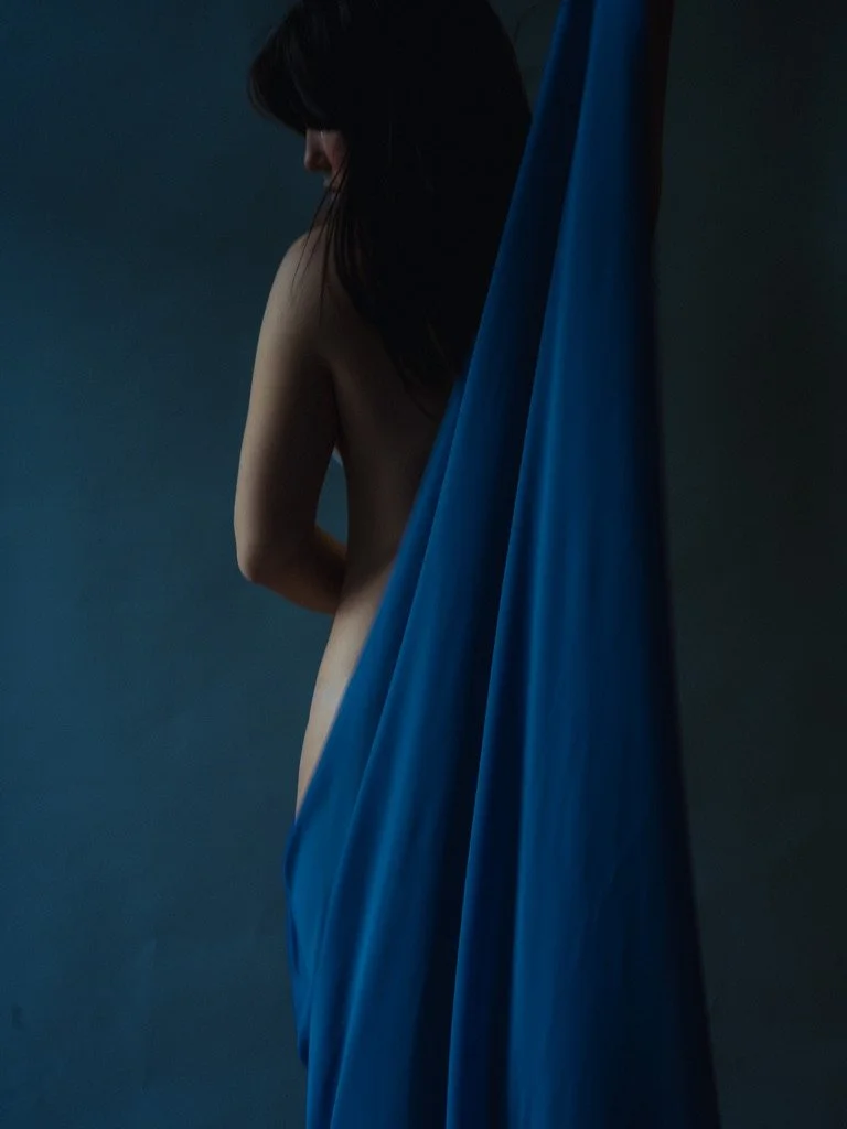 A woman standing behind a blue fabric drape, with her back partially exposed, dark hair and shadowed features.