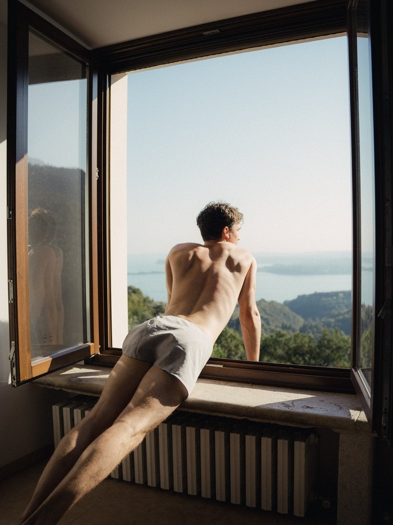 A shirtless man in white shorts leaning out of an open window overlooking a scenic landscape with water and mountains.
