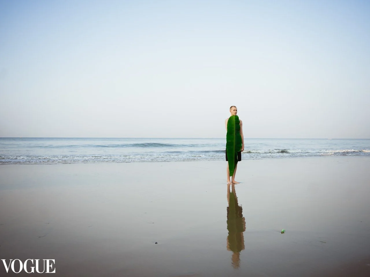 A woman standing on the beach with her reflection visible on the wet sand, holding a large green leaf in front of her body as a creative cover-up. The sky and ocean are in the background, with calm waves and a clear horizon. The VOGUE logo is in the bottom left corner.