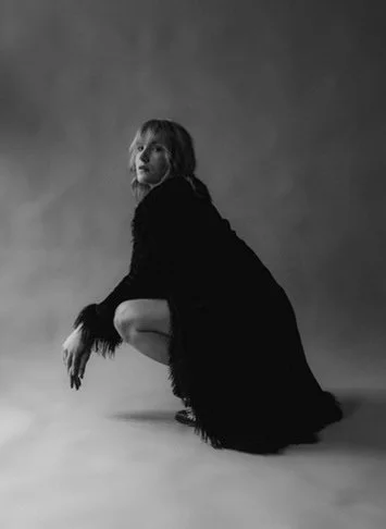 A woman with shoulder-length hair crouching on the floor in a black dress, against a plain background.