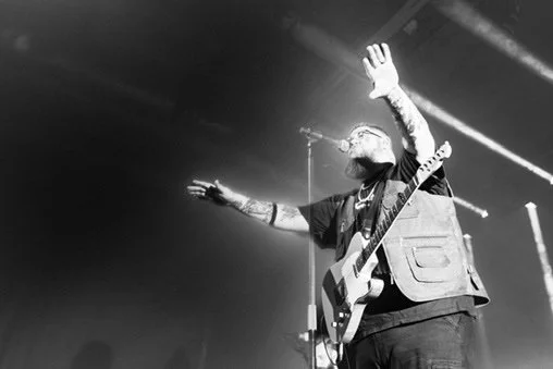 Musical performer on stage, holding a guitar with arms raised, in a black-and-white photo with stage lights overhead.