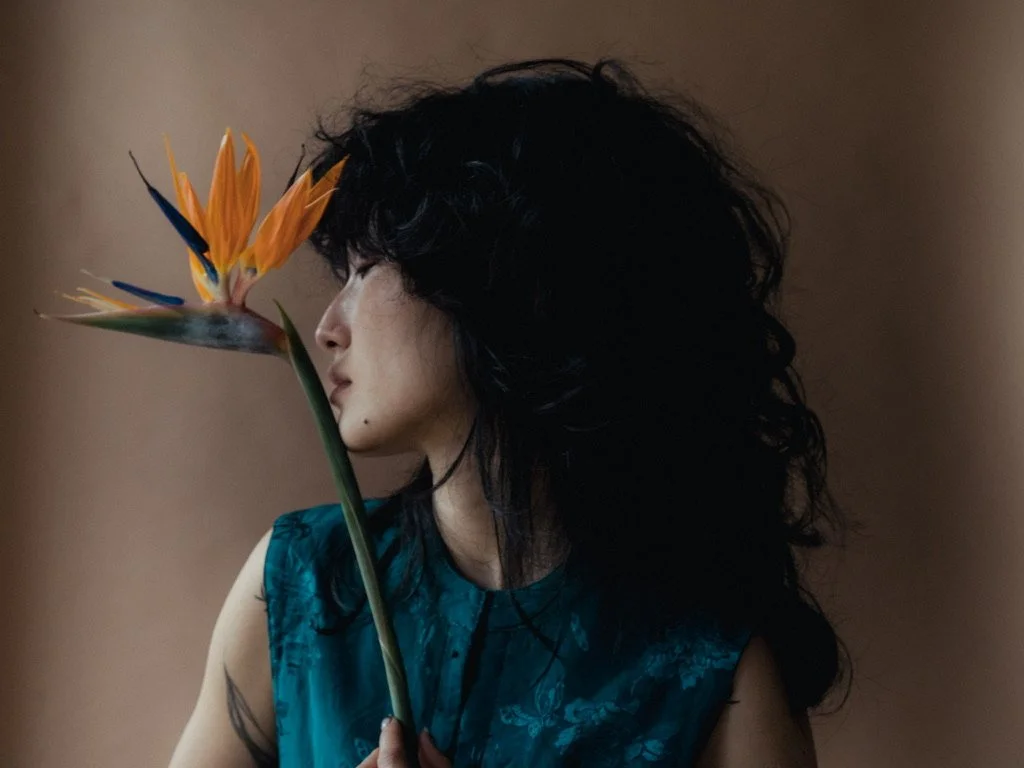 A woman with long, curly black hair wearing a blue dress holding a bird of paradise flower in front of her face, with a neutral brown background.