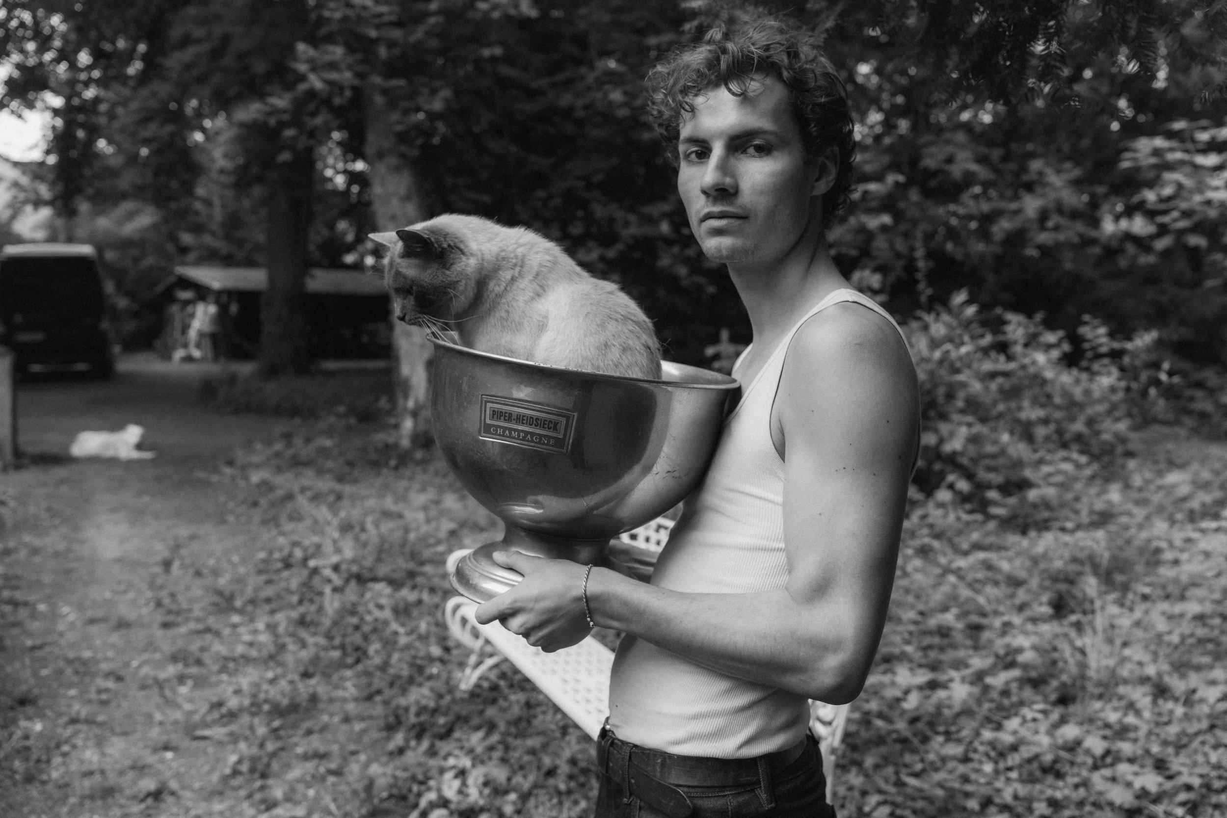 A young woman with curly hair holding a large bowl with a Siamese cat inside, outdoors in a wooded area.