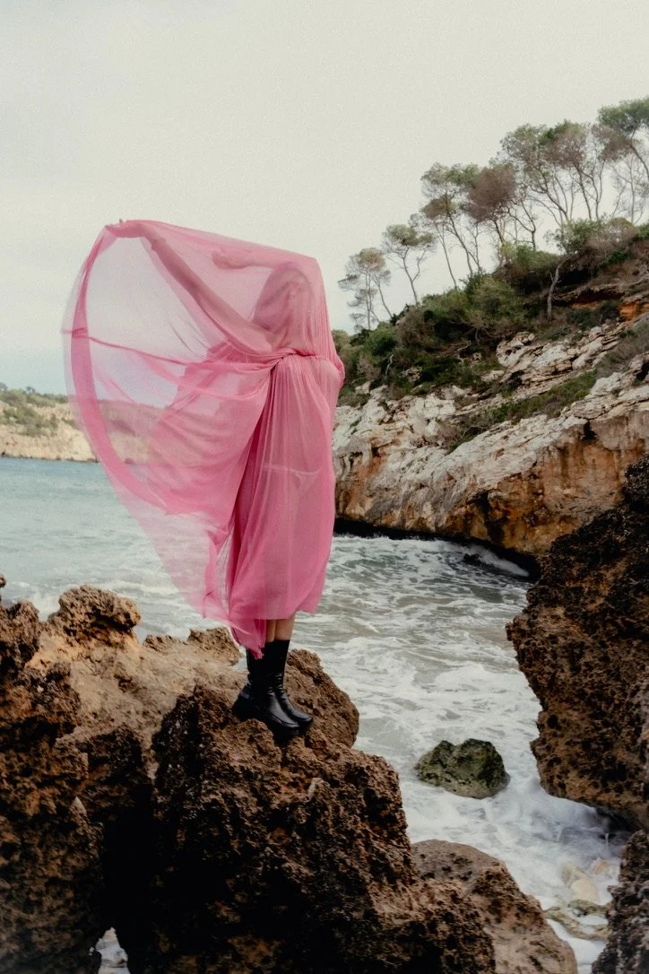 A person standing on rocks at the edge of the ocean, wearing black boots and a flowing pink dress, holding the dress up with their face and upper body obscured by the fabric, near a rocky shoreline with trees in the background.