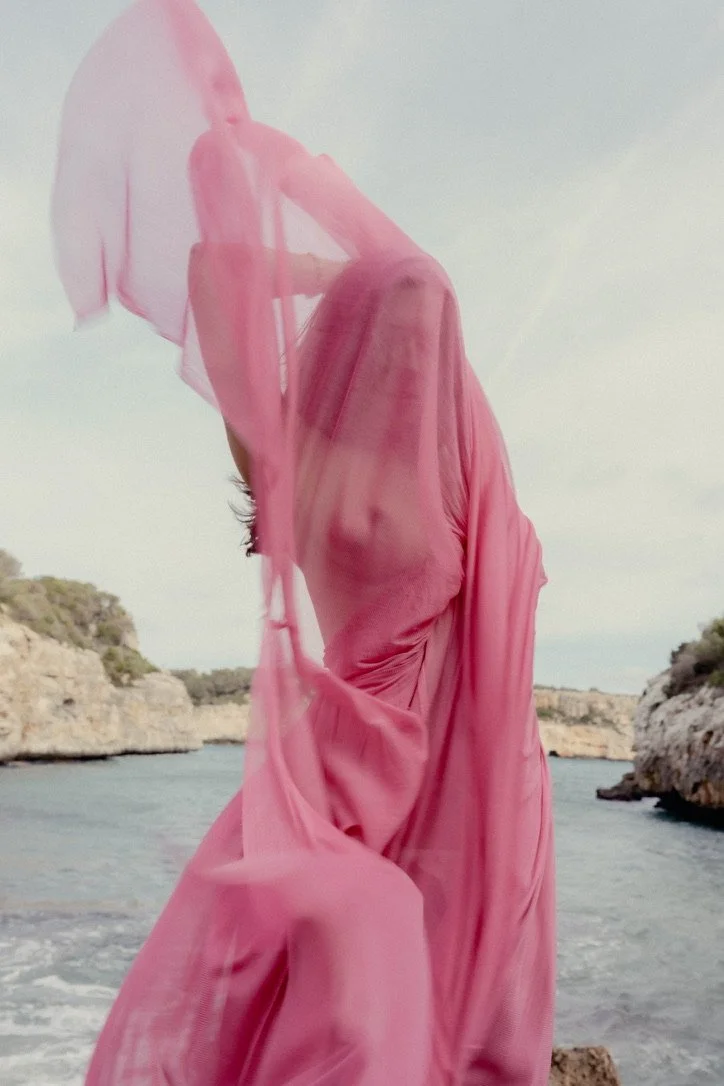 A person wearing a pink sheer fabric draped over their head and body standing near a body of water with rocky cliffs in the background.