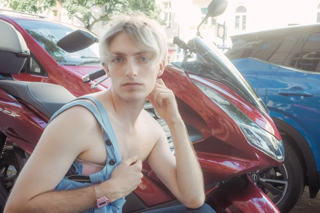 Person with light-colored hair sitting next to a red motorcycle and blue cars on a street.