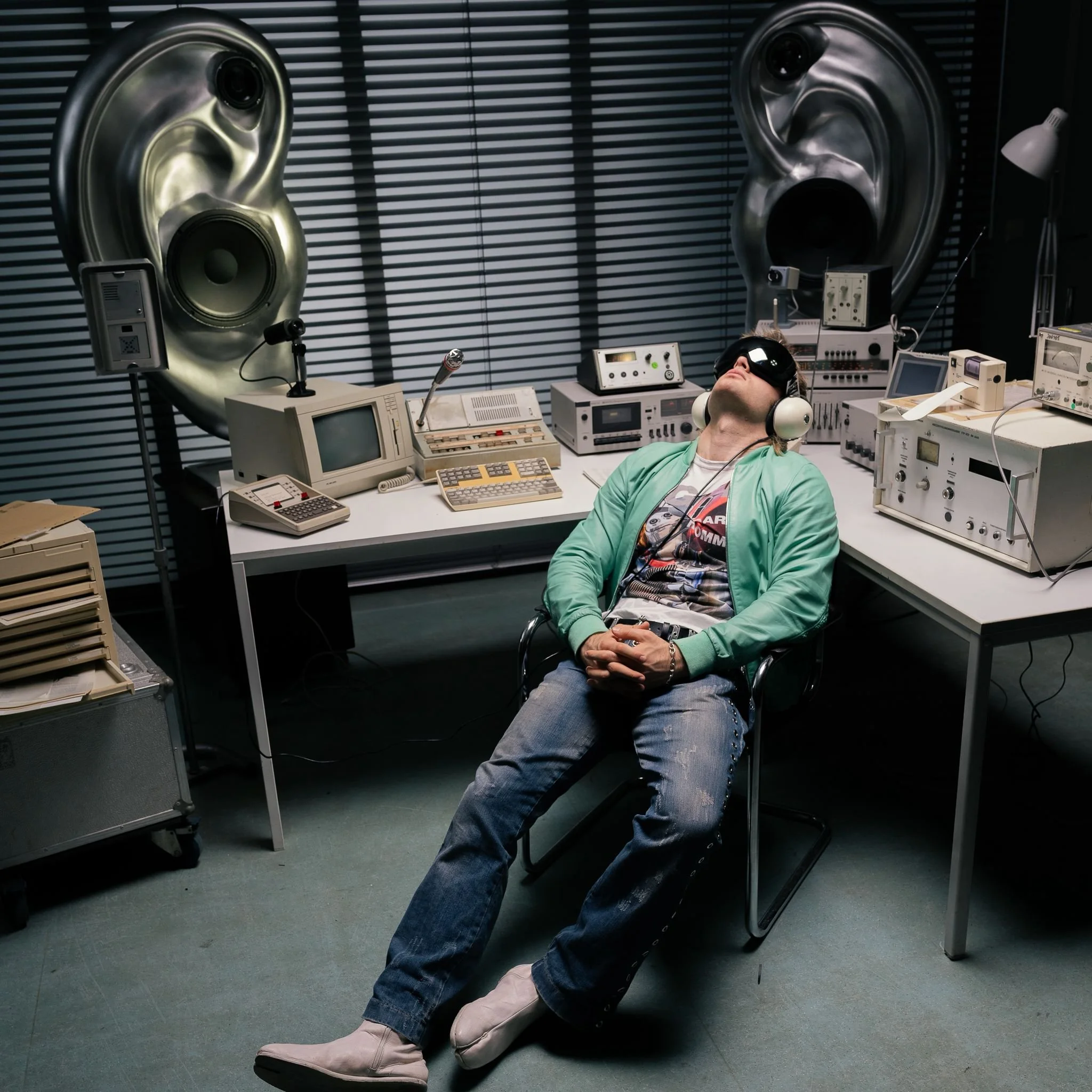 Person wearing virtual reality goggles, seated in a room filled with electronic equipment and large metallic speakers.