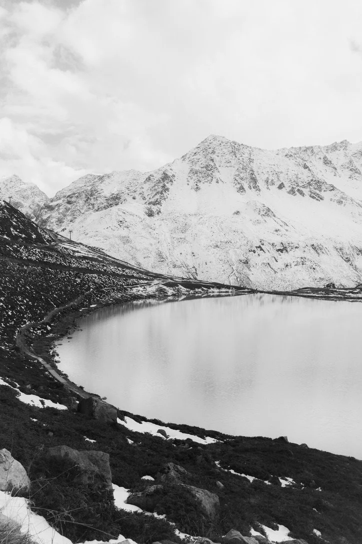 Black-and-white photo of a mountain landscape with a snow-capped mountain range, a lake in the foreground, and snow patches on the surrounding terrain.