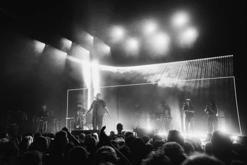 A black-and-white photo of a concert stage with musicians performing, audience in the foreground, bright stage lights shining down.