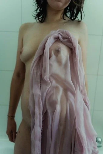 A person with dark, shoulder-length hair standing in front of a tiled wall, holding a pink sheer fabric over their chest.