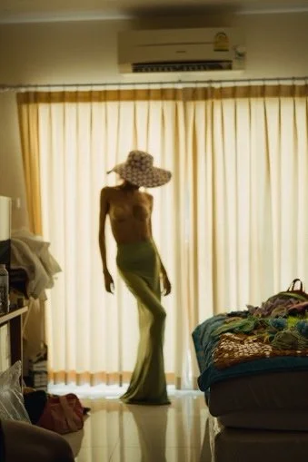 A person standing in front of closed curtains, wearing a wide-brimmed hat and loose pants.