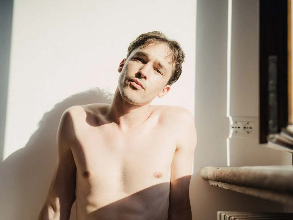 A shirtless man with short brown hair looking at the camera in a bright room with sunlight casting shadows on his face and body.