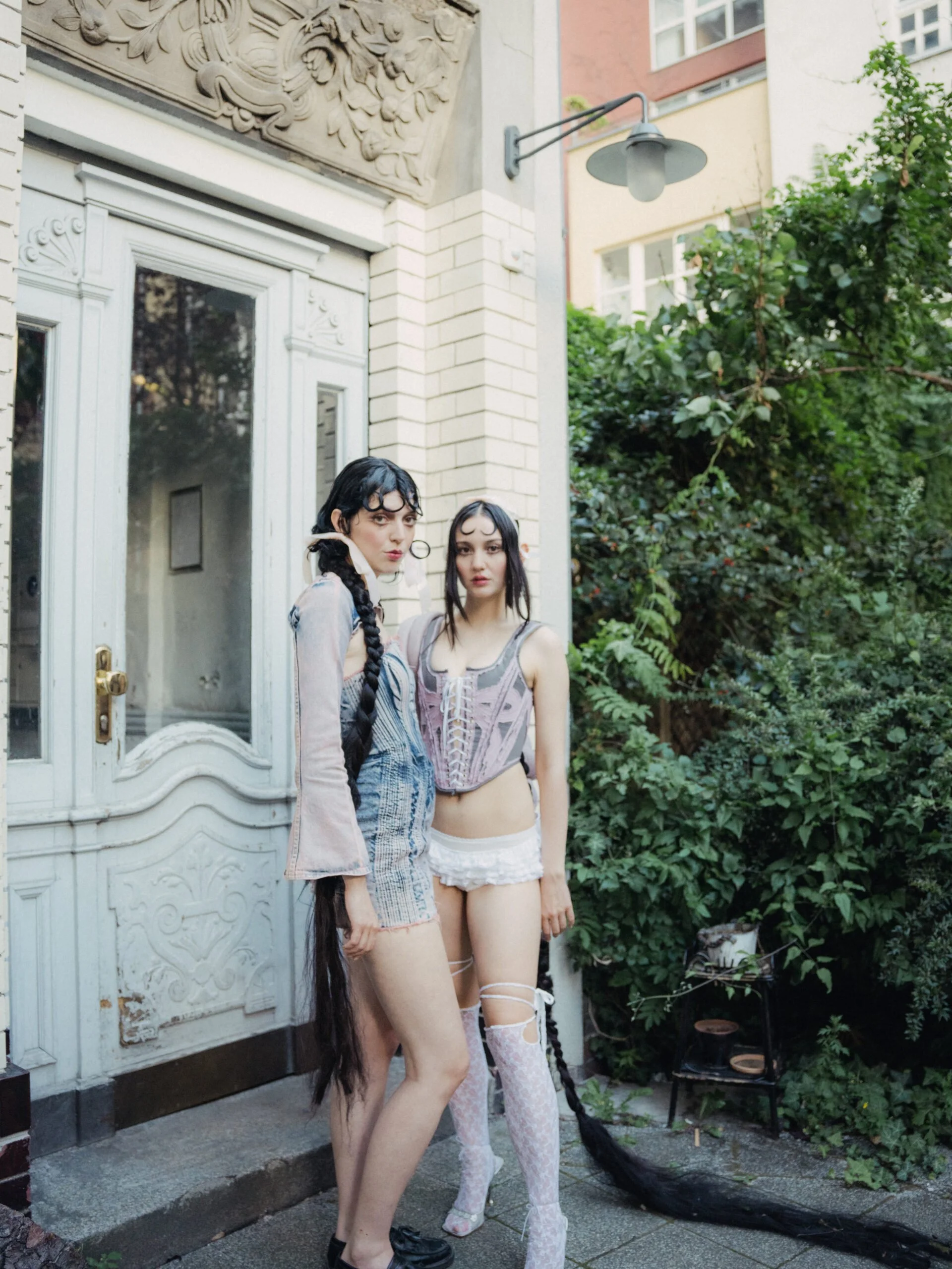 Two young women dressed in gothic and fantasy style clothing with long, braided black hair are standing outside a building. One has a shorter dress with lace-up corset and lace stockings, while the other wears a sheer long-sleeved jacket and patterned skirt. They are positioned in front of a white door with decorative trim, next to a lush green bush. A black tail is visible behind them, likely an accessory or costume piece.