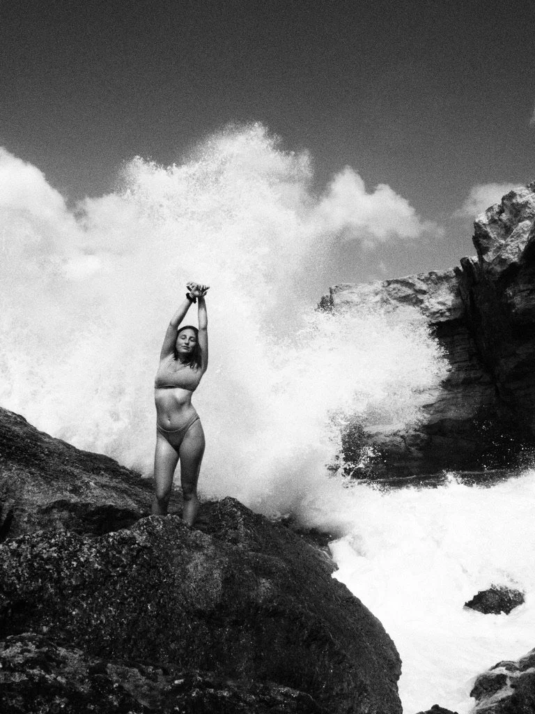 A woman in a bikini standing on rocks near the ocean with waves crashing behind her, holding her arms above her head.