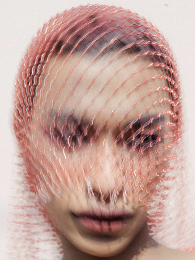 A blurred close-up of a woman's face with makeup, covered by a netted fabric with orange and pink hues, creating a distorted visual effect.