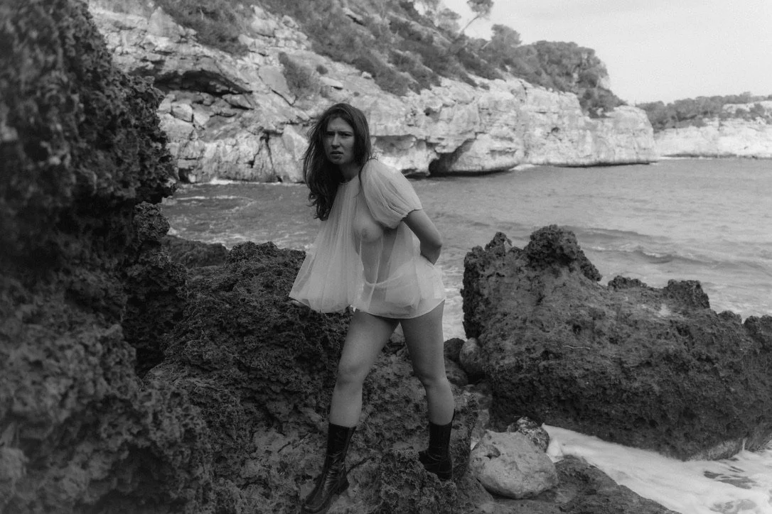 A woman standing on rocky shoreline by the ocean, wearing a sheer, short dress and boots, with cliffs in the background.